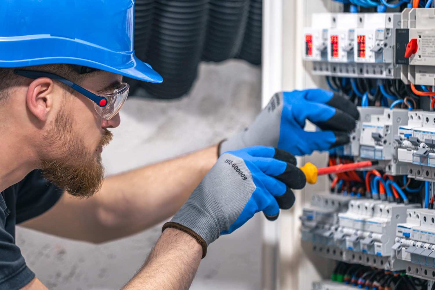 Electrician wearing high-grip work gloves using insulated screwdriver on electrical control panel – safety and precision gear in use.